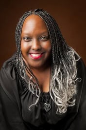 Book Review & Guest Post | Fabricated by Nicole Givens Kurtz (Cybil Lewis SF Mystery #4) 12 Author Nicole Givens Kurtz smiling warmly against a brown background. She has long braided hair with silver highlights at the ends and wears a black blouse with a heart-shaped pendant necklace.