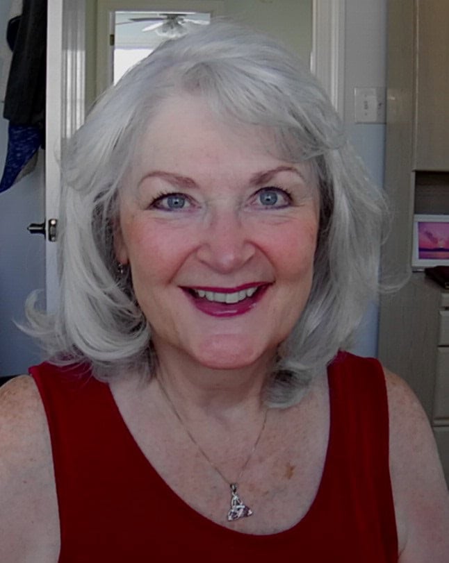 The Misremembered Lighthouse (Hayley Hunter Mystery, Book 2) by p.m. terrell | Book Review & Author Guest Post 10 Portrait of author p.m. terrell. She is smiling and wearing a sleeveless red top and silver necklace with a Celtic pendant, set against a softly lit indoor background. Used for her author bio section in the The Misremembered Lighthouse review and guest post on GinaRaeMitchell.com.