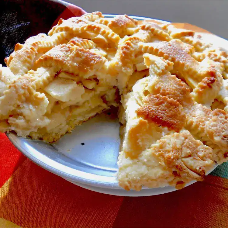 Homemade lattice-top apple pie with a golden, flaky crust and thick layers of tender apples inside. A slice has been removed to reveal the filling. The pie sits on a colorful plaid tablecloth, evoking cozy autumn baking vibes.