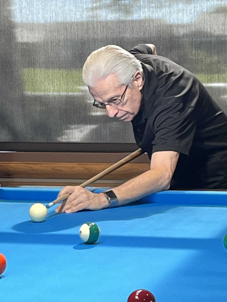 Review: Every Killer Leaves a Trace by Dennis Carson | WildBlue Press True Crime 5 Author Dennis Carson playing billiards. He leans over a blue pool table lining up a shot, wearing glasses and a black shirt. Natural light filters through a curtain in the background.