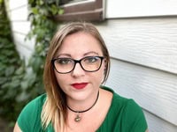 Author Kaitlin Corvus, a woman with shoulder-length blonde hair, wearing glasses and a green top, poses outdoors beside a light-colored wall and leafy vines. She has bright red lipstick and a calm expression, reflecting her creative and thoughtful personality as the author of “Nighthawks.”