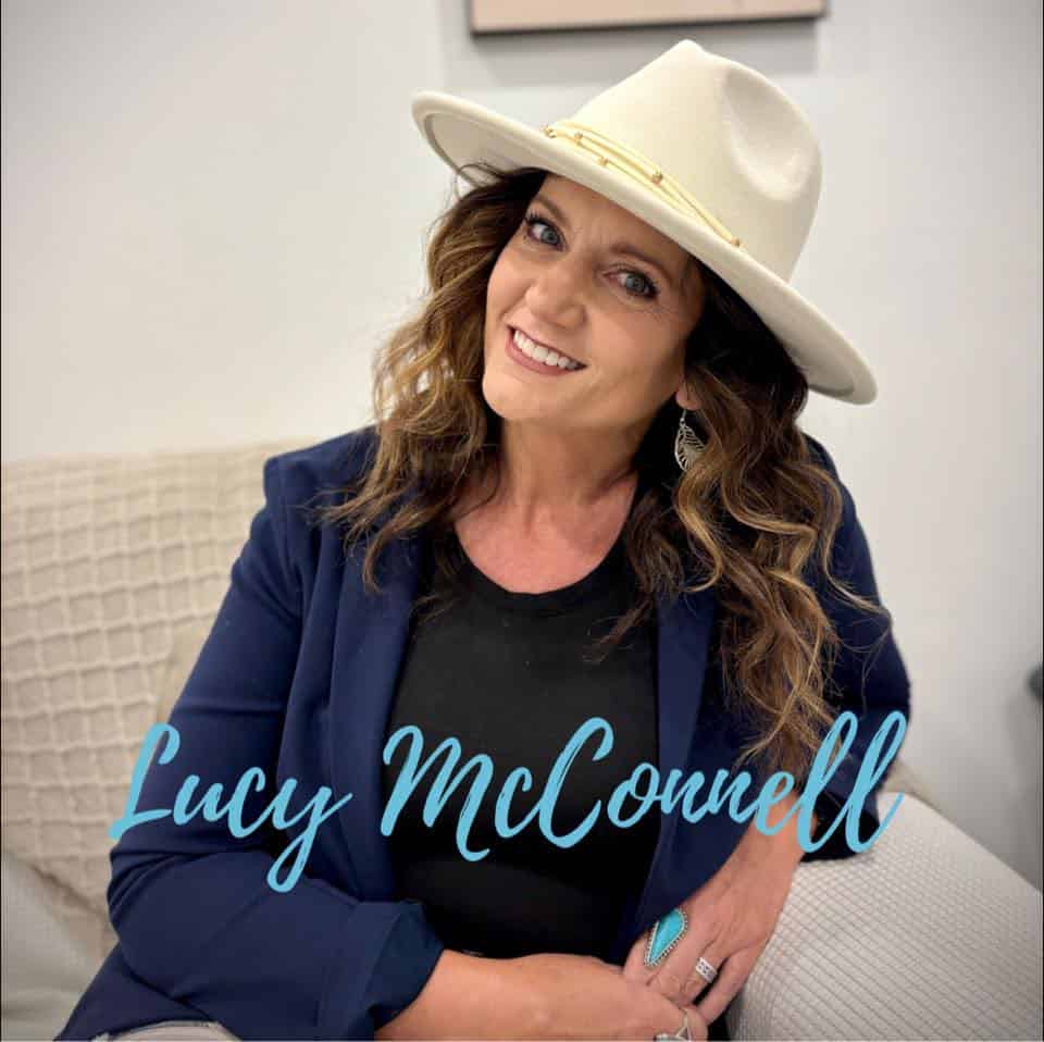 Author Lucy McConnell smiles warmly while seated indoors, wearing a cream felt hat, navy blazer, and black top with turquoise jewelry. Her name appears in light blue script. Promotional headshot used for her Christmas romance Tyler (Angel Institute #1) on ginaraemitchell.com.