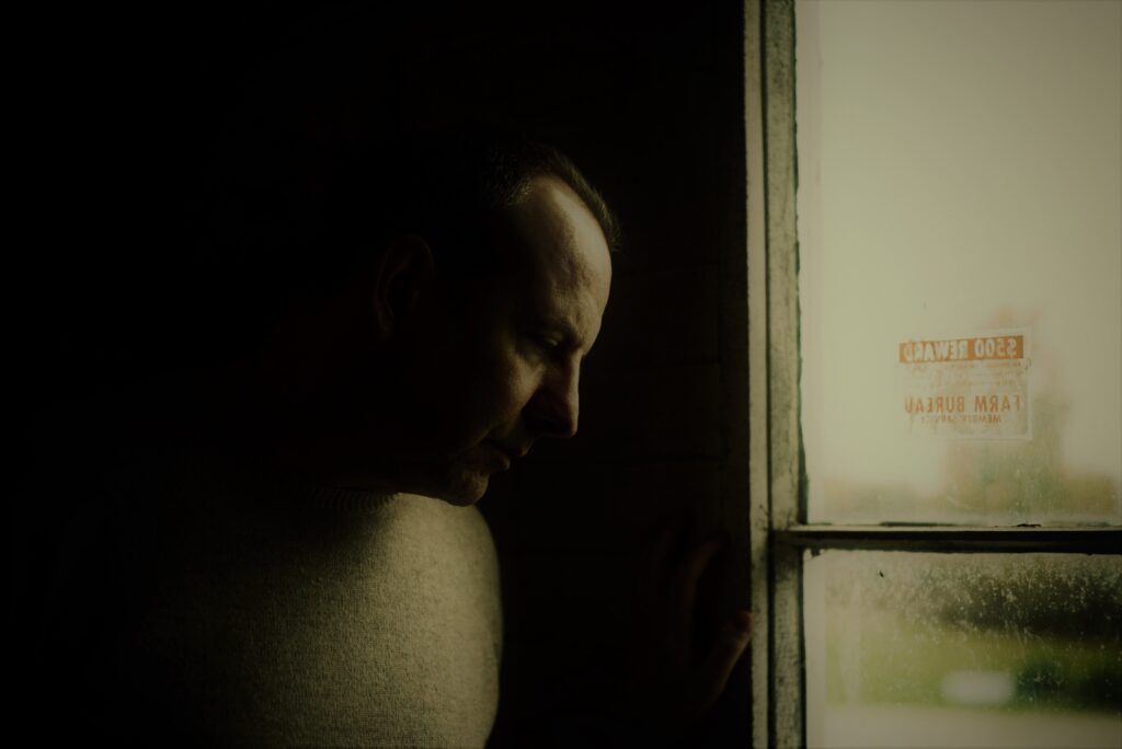 Book Review: The Nameless Land by M. Laszlo | Review, Excerpt, and Giveaway 12 Moody portrait of author M. Laszlo standing in low light beside an old window, looking downward with a contemplative expression.