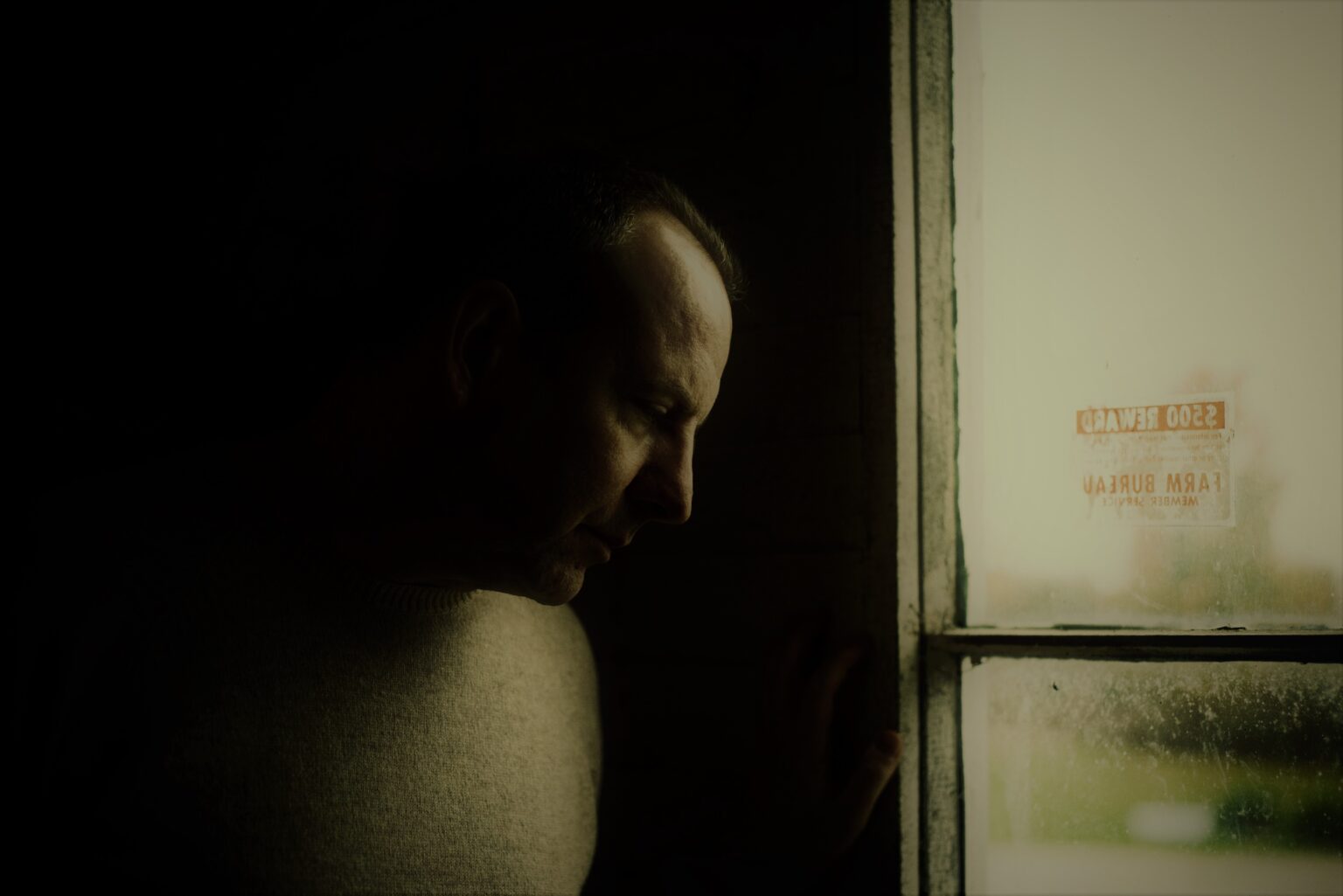 Moody portrait of author M. Laszlo standing in low light beside an old window, looking downward with a contemplative expression.