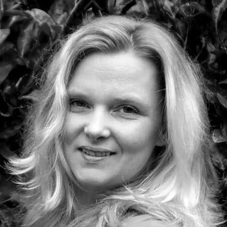 Black-and-white headshot of author Marisa Noelle smiling warmly, with shoulder-length blonde hair and leafy foliage in the background.