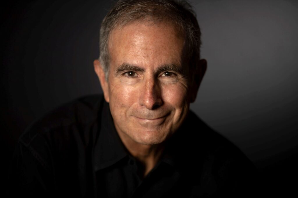 Professional headshot of author Mark Mustian. He is smiling slightly, wearing a black collared shirt, with soft lighting highlighting his face against a dark, blurred background.