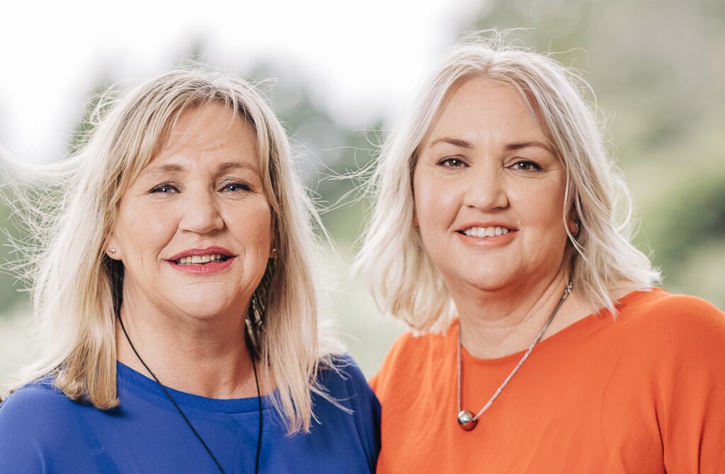 Author photo of sister writing duo Nikki Perry and Kirsty Roby. Both women are smiling at the camera outdoors with soft greenery in the background—one wearing a royal-blue top, the other an orange top. Promotional image for their novel Mary and Bright.”