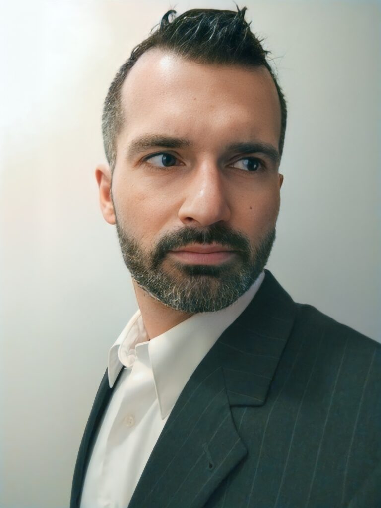 Author photo of Wes Verde. A man with short dark hair, trimmed beard, and light eyes wearing a dark suit jacket and white collared shirt, looking slightly to the side against a soft neutral background.