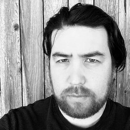 Black and white portrait of author Jon McConnell standing against a rustic wooden fence. He has dark hair, a beard, and a serious expression, wearing a dark shirt.