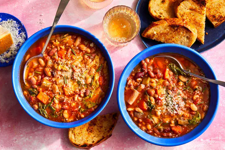 Two blue bowls filled with hearty vegetarian 15-bean soup, brimming with tomatoes, carrots, greens, and beans, topped with grated cheese. Slices of toasted bread and a glass of golden drink sit nearby on a pink tabletop, creating a warm, rustic presentation.