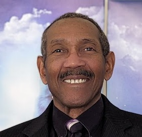 Author portrait of Austin S. Camacho, smiling in a dark suit and tie against a soft sky background. Author of the thriller novel "True Target."