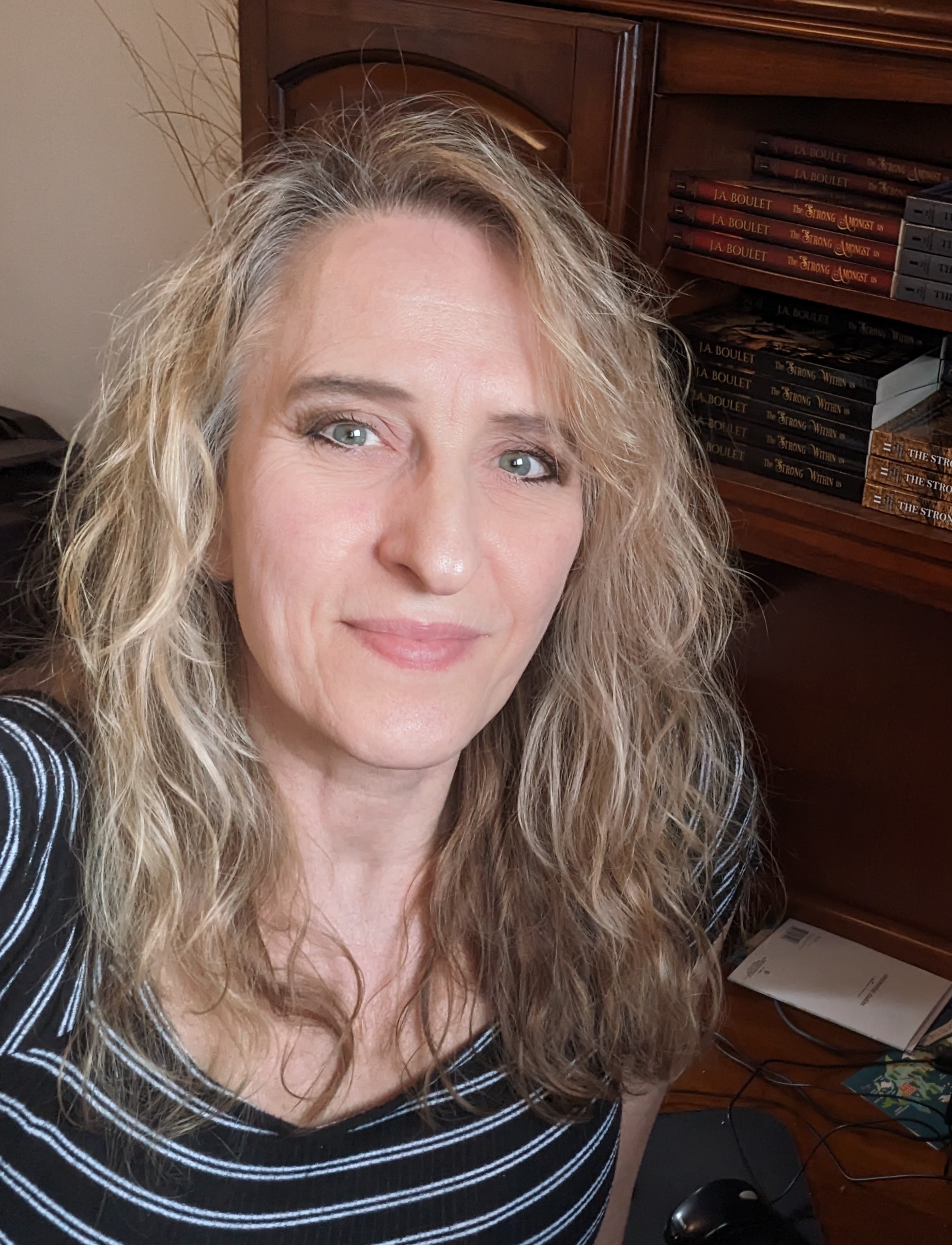 Author portrait of J.A. Boulet, historical fiction novelist, seated indoors with a bookshelf of her novels behind her. She is smiling slightly and wearing a striped top in a warmly lit home office setting.