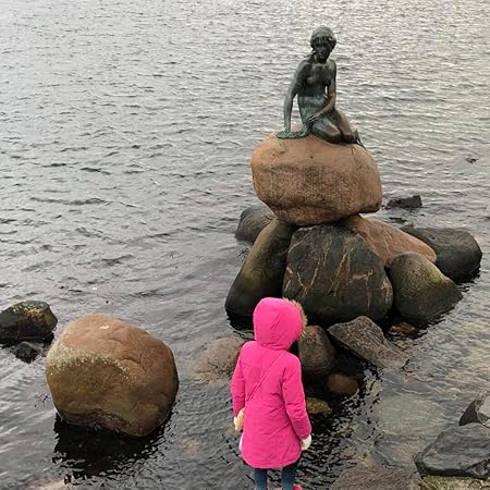 Author photo of Mescal, also known as M.S. Tanager, author of The Illumination: Izol. Shows a child standing outdoors near water with the Little Mermaid statue in Copenhagen, an image connected to the inspiration behind the Izol series.