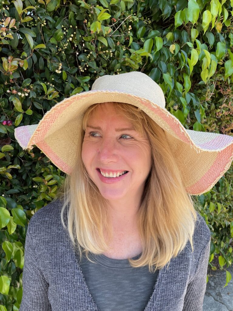 Author photo of Mary Frances Hill smiling outdoors in natural light, wearing a wide-brimmed hat and casual clothing, standing in front of green foliage.