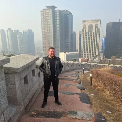 Author photo of Brian Sherlock standing outdoors on a stone walkway with a city skyline in the background. He is wearing a black jacket and dark clothing, facing the camera in a relaxed pose.