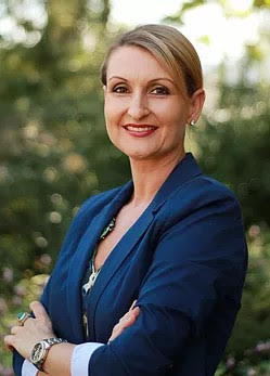 Author J.M. Hofer standing outdoors, smiling with arms crossed, wearing a navy blazer over a light top, with greenery softly blurred in the background.