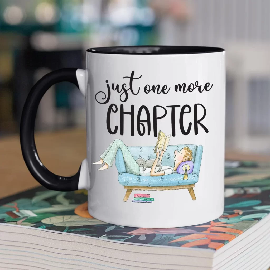 White ceramic coffee mug with black handle reading “just one more chapter,” illustrated with a cozy scene of a reader lounging on a blue couch with a book and a cat, resting on top of a stacked book.
