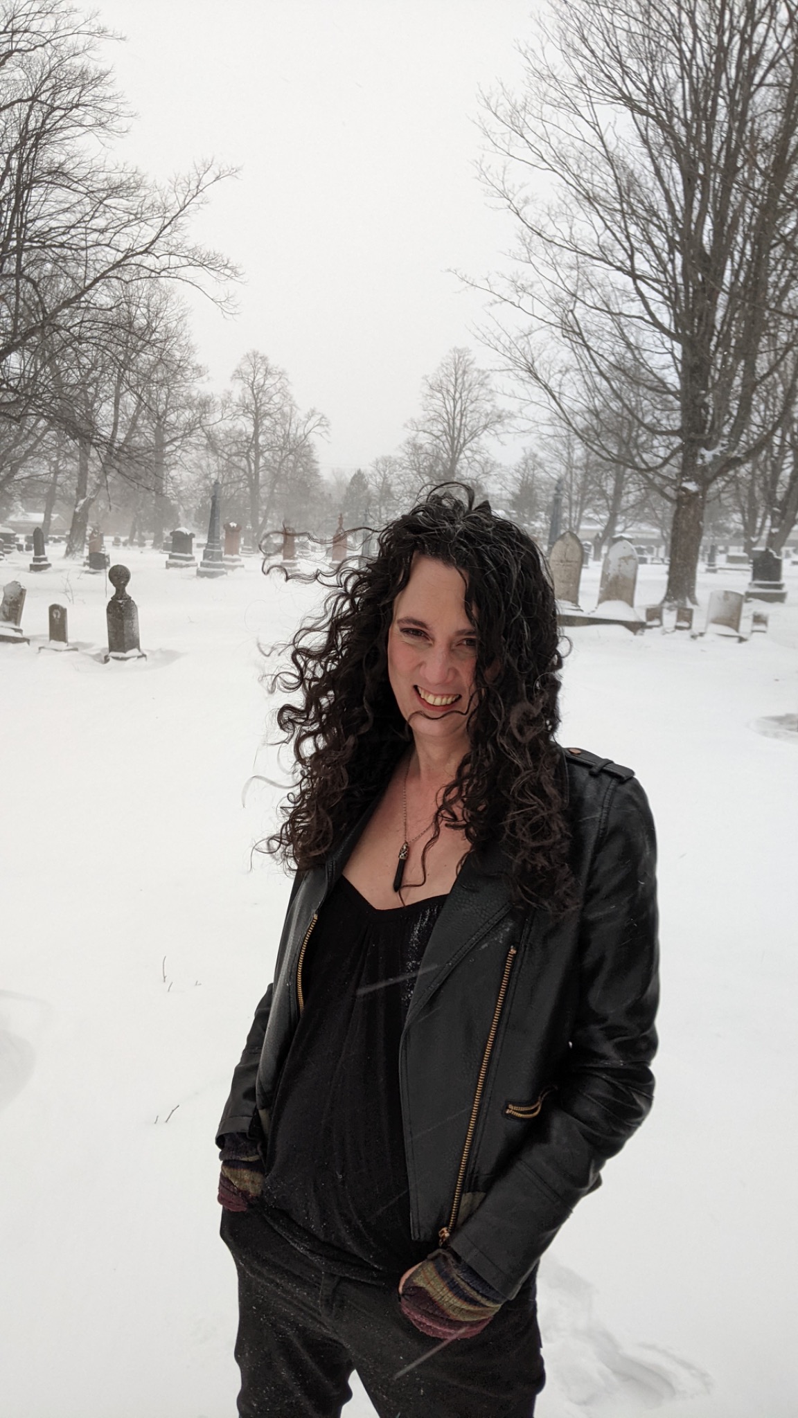 Author Leslie Allen standing outdoors in a snowy cemetery, wearing a black jacket and smiling, with long curly hair and winter trees in the background.