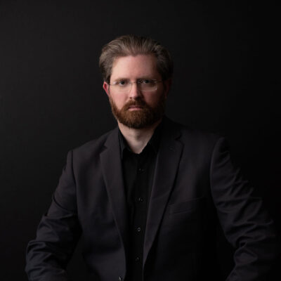 Author portrait of Seth W. James wearing glasses and a dark jacket, seated against a black background with soft studio lighting.