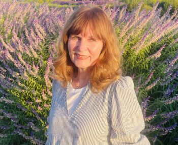 Author Bobbie Candas smiling outdoors in warm evening light, standing in front of purple flowering plants.