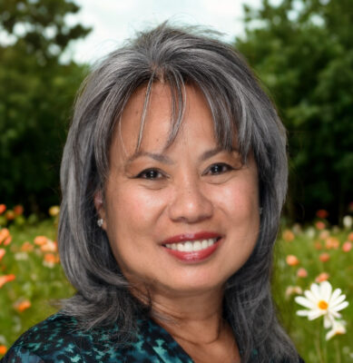 Author portrait of GG Calpo smiling outdoors with gray shoulder-length hair, wearing a dark patterned top, photographed against a softly blurred garden background with greenery and flowers.