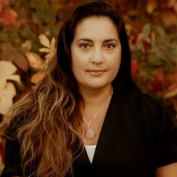 Author photo of R.A. Clarke wearing a dark top and necklace, standing outdoors against a softly blurred autumn background.