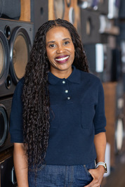 Author photo of L.N. Bokete smiling in a navy top, standing indoors beside large speakers.