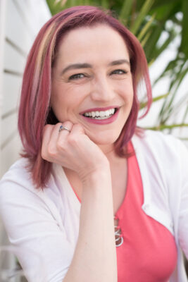 Author portrait of Rebecca Enzor smiling outdoors with pink hair, coral top, and light cardigan, photographed against soft greenery.