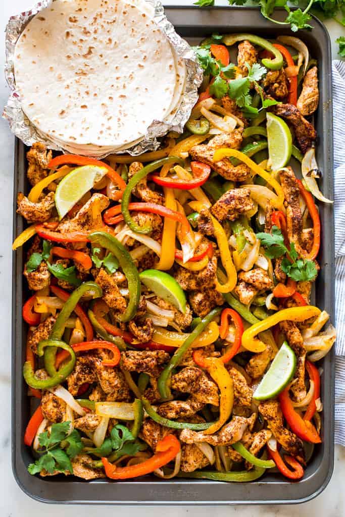 Sheet pan chicken fajitas with chicken, peppers, and onions, with flour tortillas on the side.