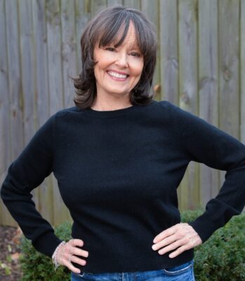 Author Susan Elizabeth Bagby smiling outdoors, wearing a black sweater, standing with hands on hips in front of a wooden fence.