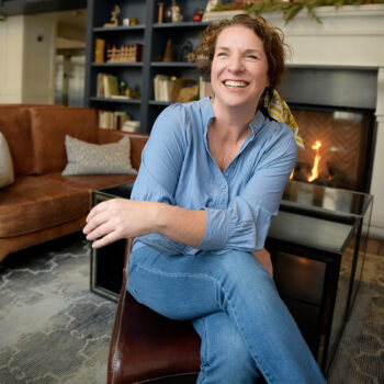 Author Kirsten Weiss seated indoors near a fireplace, smiling and wearing a blue shirt, photographed in a cozy home setting reflecting the warm tone of her humorous mystery writing.
