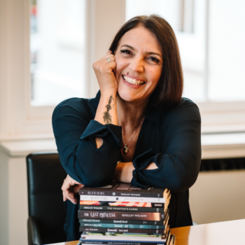 Author photo of Shelley Wilson smiling while leaning on a stack of her books at a table, wearing a dark top in a softly lit indoor setting.