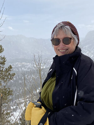Author photo of Meg Benjamin smiling outdoors in a snowy mountain setting, wearing a winter jacket, knit hat, and sunglasses.