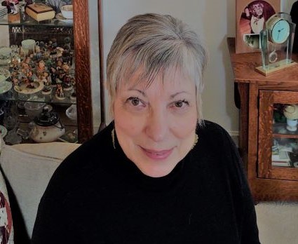 Author Merida Johns smiling in a black sweater beside a cabinet display in a warmly decorated home setting.
