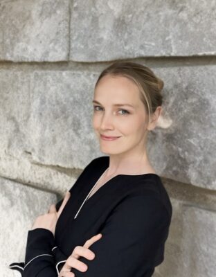 Author photo of thriller writer S.T. Ashman standing against a stone wall wearing a black blouse with white trim and smiling softly toward the camera.