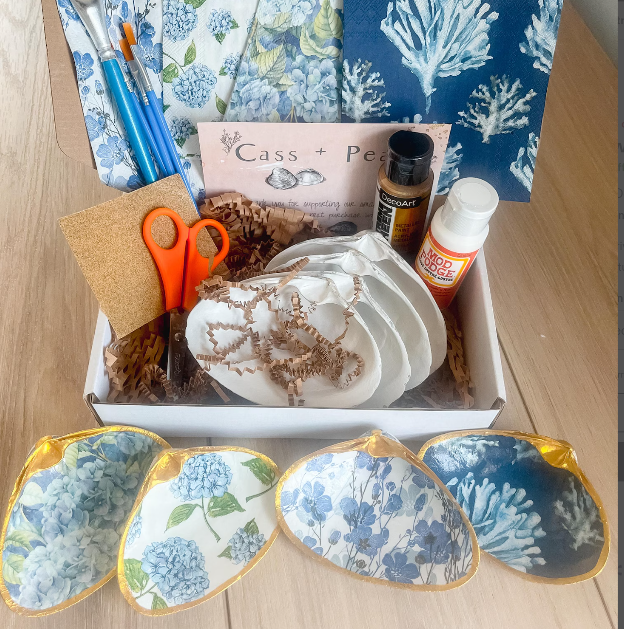 Coastal decoupage clam shell craft kit with blue floral patterns, paintbrushes, shells, scissors, and supplies arranged in a calming flat lay.
