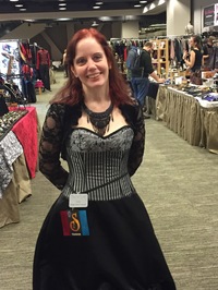Author Nikki McCormack standing at a convention or book event, wearing a black corset-style dress with lace sleeves and a conference badge. She smiles while posing in an exhibit hall surrounded by vendor booths.