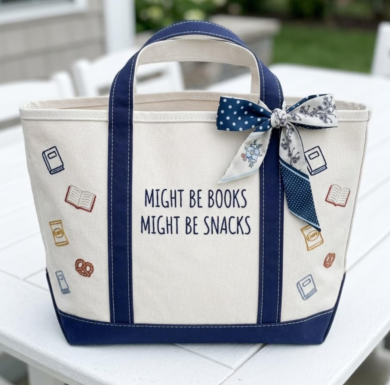 Cream canvas tote bag with navy trim embroidered with the phrase Might Be Books Might Be Snacks and decorated with small book and snack illustrations.