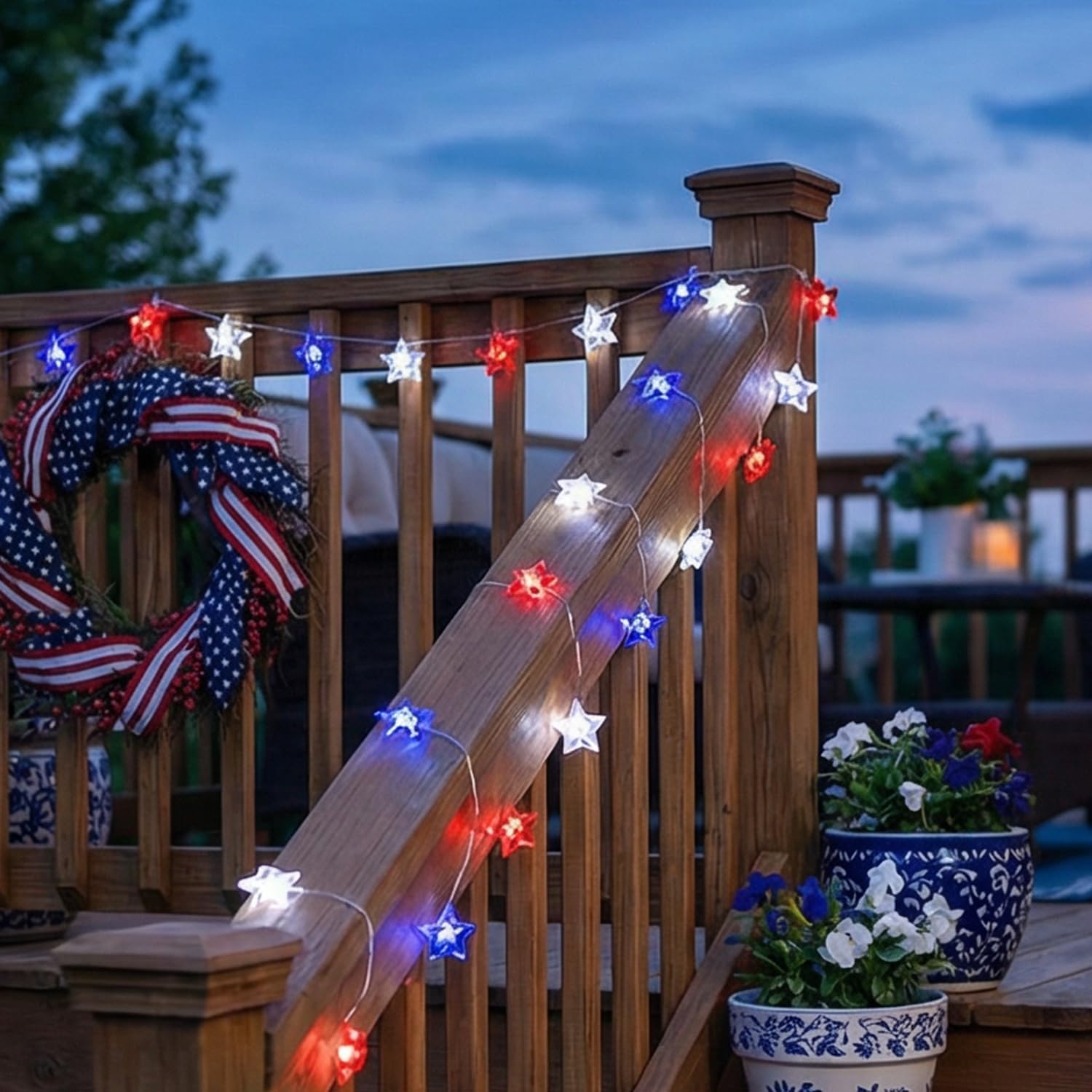 Red, white, and blue star-shaped string lights decorate a backyard deck railing at dusk beside patriotic wreaths and flower pots.