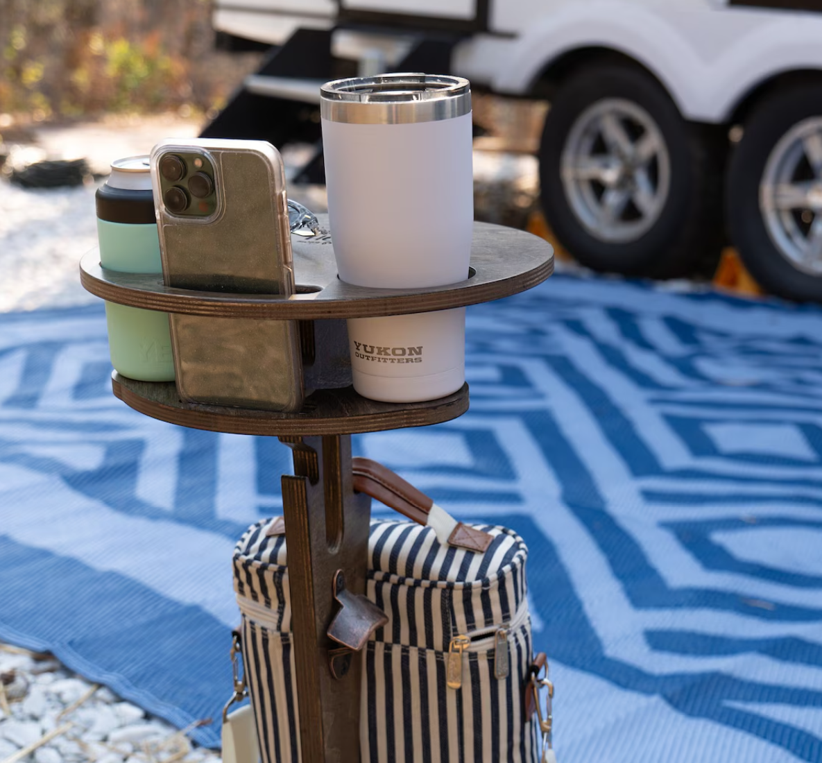 Portable wooden outdoor side table holding drinks and a smartphone beside a picnic blanket and camping setup.