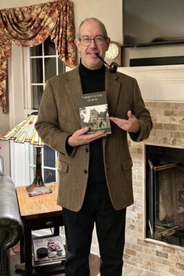 Taken to the Grave by Robert Hoffman | A Thoughtful Family Mystery About Truth and Identity 2 Author Robert Hoffman stands in a living room wearing a tweed sport coat and black turtleneck while holding a copy of Taken to the Grave and pointing toward the book cover.