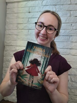 Author Kate DeMaio smiling while holding a copy of her novel, Fiona and the Forgotten Piano.