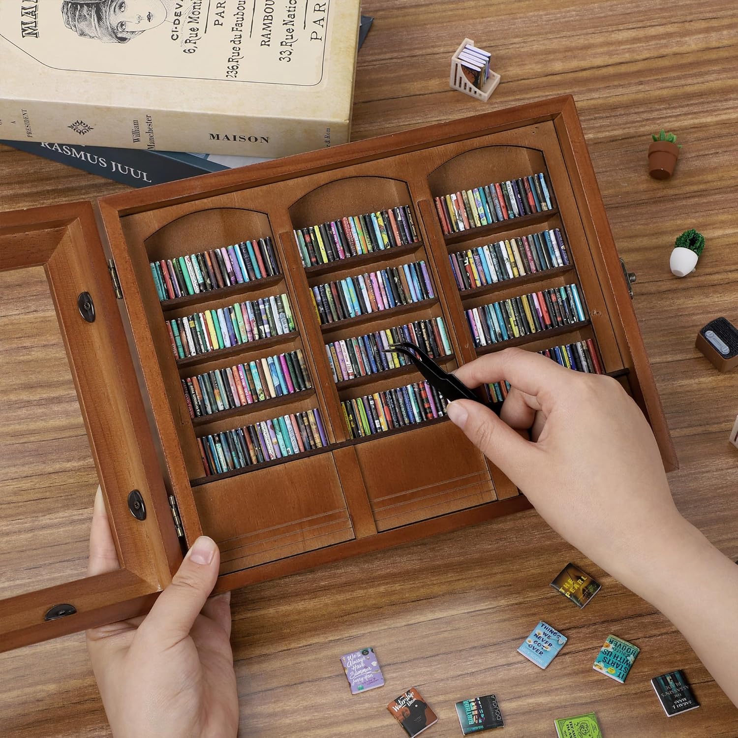Hands arranging tiny books with tweezers on a miniature wooden bookshelf